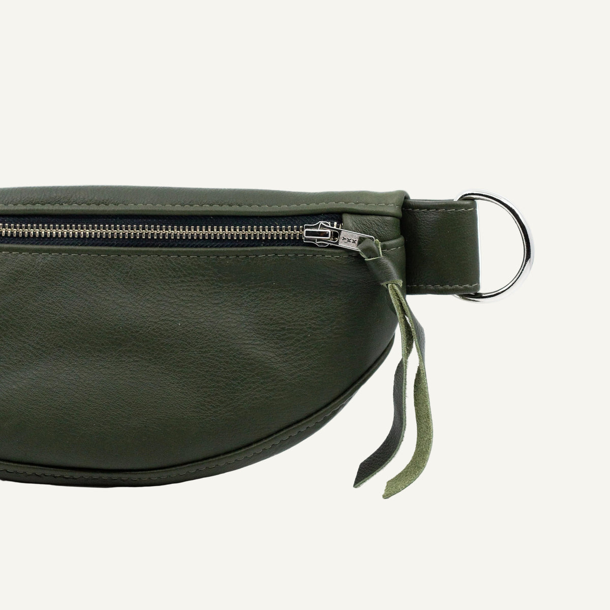 An olive green leather belt bag shown in a close-up side view, featuring a silver metal zipper, a silver D-ring, and a matching leather pull tab on a white background.
