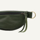 Close-up of an olive green faux-leather belt bag with a top zipper, a hanging green fabric pull, and a brass-tone metal ring on a white background.